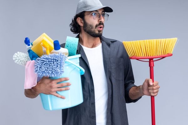 surprised looking at side young handsome cleaning guy wearing t-shirt and cap holding bucket of cleaning tools with mop isolated on white background